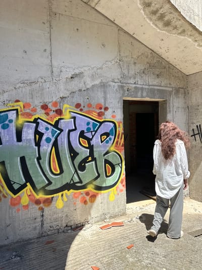 An interior view of an unfinished or abandoned concrete structure, perhaps in Gornji Katun, Montenegro. Graffiti adorns the lower portion of a wall, and a person, wearing a white shirt and gray pants, stands near an open, window-like space. The person is facing away from the viewer, their hair is brown-red.