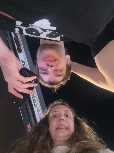 This is a low-angle selfie taken at night, featuring a man and a woman looking down towards the camera. The background is dark, indicating nighttime, with illuminated signs providing light from below.

In the lower-left portion of the frame, a man with light skin and short brown hair is visible. He wears a black t-shirt with a prominent white graphic design on the left side. He makes a playful expression, winking with his left eye and slightly sticking out his tongue. His right hand holds a dark smartphone, which is angled towards the viewer.

In the upper-right section, a woman with long, wavy brown hair and light skin is seen. She smiles broadly, revealing her teeth, and looks directly towards the camera. Both individuals appear to be in good spirits, with their faces illuminated from below, likely by the ambient light from the surrounding street signs.

Behind and above the subjects, several illuminated signs are visible. These signs display characters that appear to be Korean script, including the numbers "1999" and words resembling "슈퍼" (supermarket) and "닭갈비" (dakgalbi, a Korean dish). The setting suggests an urban environment at night.