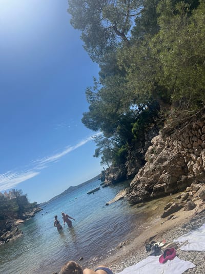 The image depicts a beach in Peguera, Spain. Two individuals are wading in the water. The beach has a rocky shoreline with trees and vegetation. Towels and personal items are on the beach. The sky is blue with a few clouds.