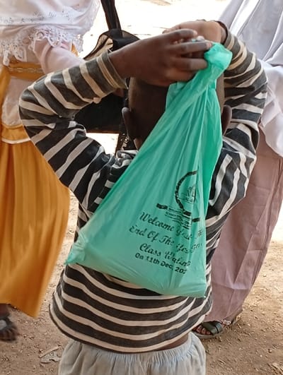 The image, taken in Fanisau, Nigeria, shows a young child from behind, wearing a grey and white striped long-sleeved shirt and light-colored shorts. The child is holding a light green plastic bag with both hands, raised towards the back of their head.
Partial views of other individuals are visible around the child. To the left, a person is dressed in a white top with lace trim over a yellow or orange skirt. To the right, another person wears a white garment and what appears to be a light brown or purple lower garment, with a foot visible wearing a dark sandal.
The scene is set outdoors on a dirt ground, under bright, clear daylight conditions, suggesting a sunny day. The activity appears to be part of a gathering or event, possibly where items are being distributed or received.
A notable detail is the text printed in black on the light green plastic bag. It includes a logo at the top and the following words:
"Welcome Your"
"End Of The Year" (interpreting "Ifo" as "Year")
"Class Walimah" (interpreting "Waling" as "Walimah," an Islamic celebratory gathering)
"On 11th Dec, 2023" (interpreting "202" as "2023")
This text indicates the bag is likely a party favor or gift from an "End of the Year Class Walimah" held on December 11th, 2023. The overall impression is that of a community or school celebration.