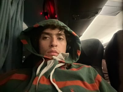 An upside-down close-up shot features a person wearing a green hoodie with orange stripes and white drawstrings. Their face is visible within the hood, gazing upward. The background shows airplane seats, with black headrests and a dark interior. A glimpse of a window and some light is visible.