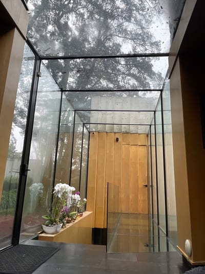 The image displays a modern interior architectural space, likely an atrium or hallway, located in Fraccionamiento y Club de Golf los Encinos, Mexico. Extensive use of clear glass forms the left wall and ceiling, revealing an exterior environment where raindrops are visible on the glass surfaces.

The floor is dark gray, with a black rectangular mat positioned in the lower-left foreground. Through the glass on the left, a large, mature tree with dense foliage is visible, along with general greenery and a distant light-colored concrete or stone wall.

Along the base of the left glass wall, a light-colored ledge supports a display of multiple white and purple Phalaenopsis orchids, interspersed with small brown glass vessels.

Straight ahead, a wall constructed from vertical light brown wooden panels features a flush-mounted door with a black handle. A small black cylindrical device, likely a security camera or sensor, is mounted on this wooden wall above and to the left of the door.

To the right, a solid tan wall is visible, with a small white circular object affixed to its lower section. A clear glass balustrade extends from the floor towards the right, indicating a drop-off or a descending staircase. No individuals are present in the scene.