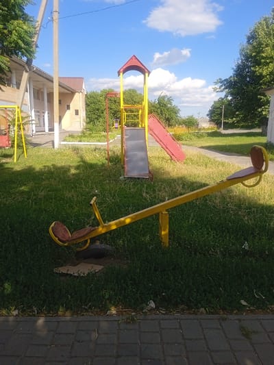 This image captures a bright, sunny day at a playground in Maksymivka, Ukraine. The scene features a seesaw in the foreground, made of yellow metal with weathered brown seats. Behind it, a red and yellow slide structure stands against a backdrop of lush green grass and trees. A building with a light-colored facade and a brown roof is visible to the left, suggesting the playground might be near a school or residential area. The sky is a vibrant blue with fluffy white clouds, indicating fair weather. The time of day appears to be late morning or early afternoon, given the strong sunlight and shadows. The overall atmosphere is peaceful and ordinary, depicting a typical day in a community park. There is no visible text in the image.