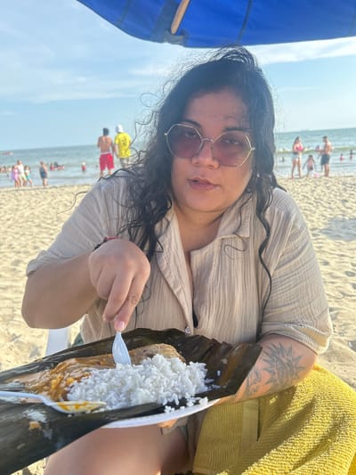 An individual is depicted seated on a sandy beach in General Villamil Playas, Ecuador, positioned beneath the partial shade of a blue umbrella. The subject, a female with dark, wavy hair, wears hexagonal-frame sunglasses with pinkish lenses and a light tan, crinkle-textured collared shirt. A tattoo, resembling a sun design, is visible on her left forearm. She holds a white plastic fork/spoon over a white disposable plate containing a meal, which rests on a dark green leaf. The plate holds a serving of white rice adjacent to a saucy, orange-brown dish. A yellow towel is draped beside her on the right. In the background, the ocean extends under a clear blue sky, populated by numerous individuals swimming and wading. Other people are visible on the sunlit beach in the mid-distance.