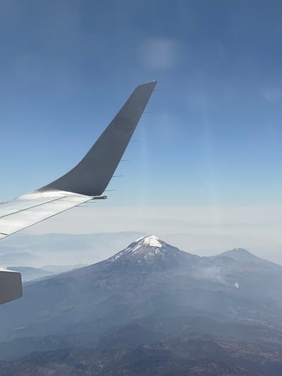 An airplane wing stretches across a blue sky, equipped with static wicks along its trailing edge. Beyond the wing, a hazy mountain range dominates the background. The foremost peak has a snow-capped summit. The image is rotated counterclockwise, giving the mountains a tilted appearance. The lighting suggests a bright, sunny day.