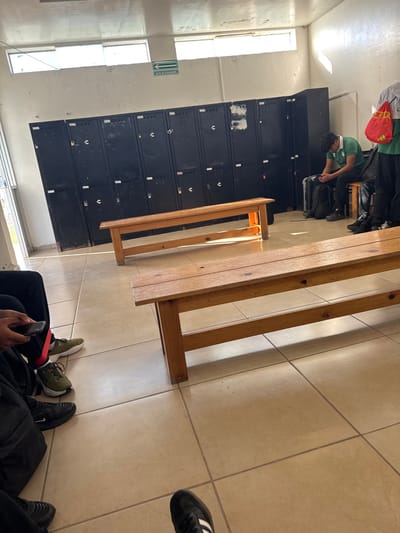 The image depicts an indoor locker room or changing facility located in La Venta, Mexico. The room features light tan tiled flooring with dark grout lines and white-painted walls, which exhibit minor scuffing and discoloration. A green "SALIDA DE EMERGENCIA" (Emergency Exit) sign is affixed to the far wall, positioned below two high-set rectangular windows that provide natural light. A full-length bank of approximately ten dark grey metal lockers with individual latches and multiple padlocks spans the far wall, some displaying wear or adhesive residue.
Two unvarnished, light-colored wooden benches run parallel to the locker bank.
Four individuals are present. On the far right, a person is partially visible, standing, wearing a green shirt, dark pants, and dark footwear, carrying a red drawstring bag with a white logo. To their immediate left, a male individual is seated on a dark stool or bench, dressed in a green polo shirt with white trim and dark pants, with a black backpack at his feet. His head is lowered, and he appears to be focused on an object held in his hands. In the left foreground, the lower half of an individual is visible, wearing black athletic pants with a red inner stripe near the ankle and olive-green athletic shoes. This individual holds a dark mobile phone in their hands. In the right foreground, the feet of a fourth person are visible, clad in black athletic shoes with white stripes. No direct interactions between the individuals are depicted.