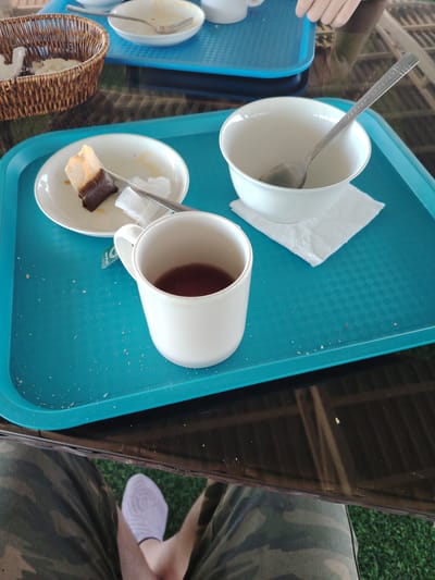 A close-up, high-angle shot shows a turquoise tray on a dark reflective surface, possibly a table. The tray contains a white mug half-filled with dark liquid, a small white plate with a used tea bag and a napkin, and an empty white bowl with a spoon resting inside. Small crumbs are scattered on the tray. In the lower part of the frame, a person's legs are visible, wearing camouflage pants and a white sock with a patterned design. The background appears to be an outdoor setting with green grass. The scene is likely set in Toranghylyk, Kazakhstan.