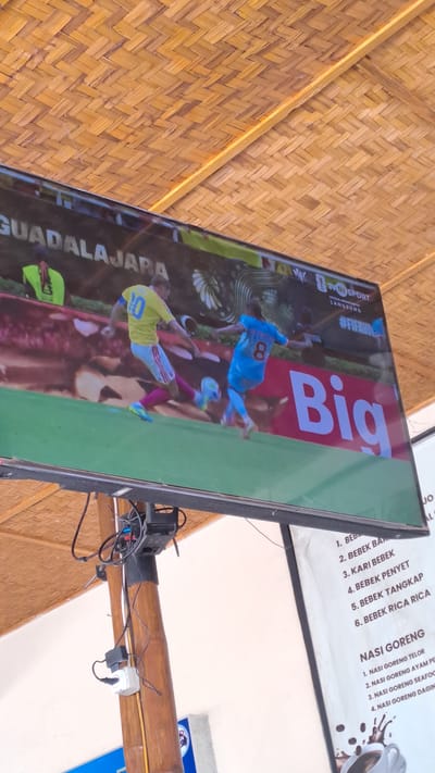 This media file shows a television screen displaying a soccer match. Two players in soccer uniforms are visible, one in a blue jersey with the number 8, and another in a yellow jersey with the number 10. The screen has the text "GUADALAJARA" and "#FIFAVI" visible. To the right of the soccer match, there is a menu board with Indonesian text listing food items such as "BEBEK" (duck) and "NASI GORENG" (fried rice). The television is mounted on a wooden beam, and the background appears to be a woven bamboo ceiling. The overall setting suggests an informal establishment, possibly a restaurant or a food stall, in Julikeude Dua, Indonesia. The lighting is ambient, and there are no clear indicators of the time of day or weather. The focus of the image is the television screen and its content, which is a sporting event.