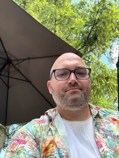 In Lozno, Bulgaria, a bald man with glasses and a beard is pictured outdoors, under a partially visible dark umbrella. He is wearing a white t-shirt and a colorful floral print shirt. The background consists of lush green trees and a bright blue sky, suggesting a sunny day. The man is looking directly at the camera, with a neutral expression. The image is taken from a close-up perspective, focusing on the man's face and upper torso, with the umbrella and natural scenery framing him.