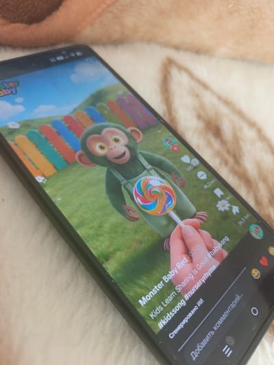 This is a close-up, angled shot of a smartphone screen displaying a video of a cartoon monkey character holding a lollipop. The monkey is green with large yellow eyes and is wearing green overalls. It is set against a backdrop of a grassy field with a colorful, picket-style fence. Small white flowers are scattered in the grass. The video appears to be a children's educational song or animation, as indicated by the text on the screen.
The screen displays several interactive elements common to video-sharing platforms. On the right side, icons for likes, comments, and shares are visible, along with numerical counts in Russian indicating their popularity (e.g., "585 тыс." which means 585 thousand). Below the video, a caption reads "Monster Baby Red . 02-26 Kids Learn Sharing Is Good #babysong #kidssong #nurseryrhyme." There's also text indicating the video was "Сгенерировано ИИ" (AI-generated). At the bottom of the screen, there is a text field prompting the user to "Добавить комментарий..." (Add a comment...).
The phone itself is angled and appears to be resting on a textured, fuzzy surface, possibly a blanket or carpet, which is a light beige or cream color. The background above the phone is slightly out of focus and shows a hint of orange fabric. The overall lighting suggests an indoor setting, and there are no clear indicators of the time of day or weather. The location context provided is Gornyak, Russia.
