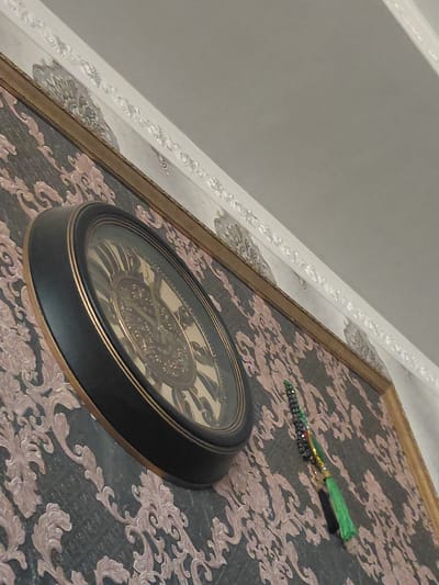 The image shows a decorative clock with a black frame and a gold-colored face. It is hanging on a wall covered with dark wallpaper featuring a light pink ornate floral pattern. Above the wallpaper, there is a decorative white trim with floral motifs. To the right of the clock, a string of dark beads with a green tassel hangs down. The scene appears to be indoors, possibly in a residential setting. The image does not contain information that can place it in Preobrazhenskoye, Russia, or any other specific location.