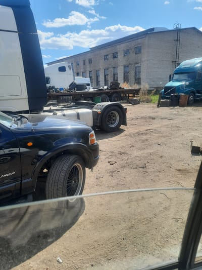 The image appears to show a yard or parking area in Научный Городок, Russia. There are several semi-trucks parked in the area. A brick building is also visible in the background, and the ground appears to be dirt or gravel. A black Ford Explorer is parked nearby.