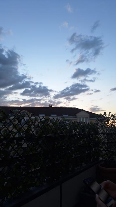 The image depicts a view from a balcony overlooking rooftops and the sky in Gournay-sur-Marne, France. The sky is a pale blue transitioning to darker hues towards the horizon, with scattered grey clouds. In the foreground, a dark green lattice fence with climbing foliage partially obscures the view of a building with a tiled roof, dormer windows, and a chimney. The building appears to be residential. Below the fence, a dark railing runs along the balcony. To the right, a hand is visible holding a smartphone, with a terracotta pot in the background. The scene is captured during dusk or dawn, as indicated by the lighting conditions.