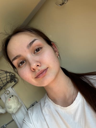 A young adult female is depicted in a close-up selfie, situated within what appears to be a train compartment, likely in or passing through the city of Kalachinsk, Russia. The subject has a light complexion, dark brown hair pulled back, and dark brown eyes, looking directly at the camera with a neutral expression. She wears a white crew-neck t-shirt. Visible accessories include a clear-stoned stud earring in her right earlobe and a small gold-toned hoop in her right upper ear cartilage. A thin, gold-toned chain necklace is visible around her neck.
The background consists of light beige wall panels. To the left, a metallic, grid-patterned overhead luggage rack is partially visible. Draped below the rack is a white terry cloth towel. Attached near the towel is a small, white, spherical plush keychain resembling a seal, featuring small black eyes and faint pink cheeks. A small, chrome-colored hook is affixed to the ceiling in the upper portion of the frame. The lighting is even, suggesting ambient indoor illumination.