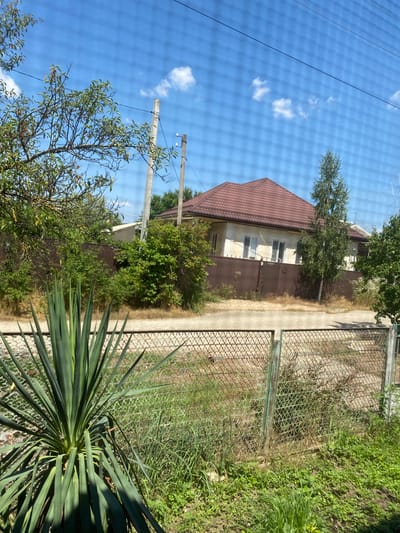 A bright, sunlit day in Akhtyrskiy, Russia, is depicted through a mesh-like overlay, suggesting the photo was taken through a fence or window screen. In the foreground, a large, spiky yucca plant with long, green leaves dominates the left side. Behind it, a wire-mesh fence runs horizontally, with patches of green grass and dry, brown vegetation growing through and around it.
Beyond the fence, a paved pathway or road is visible, leading to a brown metal fence that encloses a property. A single-story house with a brown, tiled roof and light-colored walls is partially visible behind the brown fence. The house has several windows with white frames. Several trees and dense green bushes are scattered around the property, adding to the suburban feel of the scene. Two utility poles with insulators stand on the right side of the pathway, with overhead wires stretching across the sky. The sky is a clear blue with a few scattered white clouds. The overall impression is a quiet, residential area on a warm, sunny day.