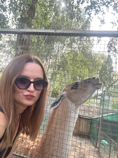 In an outdoor enclosure at a zoo in Medvedevo Urban Settlement, Russia, a woman with auburn hair and dark sunglasses poses for a selfie. She is wearing a black top and a necklace. Next to her, a llama with light brown fur peers through a metal fence, its mouth slightly open. In the background, trees with green leaves are visible, along with a structure with a corrugated roof and a green enclosure. The sun shines through the trees, creating a dappled light effect. The overall scene suggests a sunny day at the zoo.