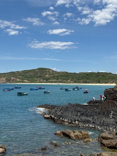 The media file displays a scenic coastal view at Phú Lương, Vietnam. The scene is dominated by a bright blue sky with scattered white clouds, suggesting fair weather during daytime. The main subjects are a row of small boats floating on clear, turquoise water. The boats are of various colors, including blue, green, and red, and appear to be fishing or leisure vessels.
In the foreground, dark, rugged volcanic rocks form a natural barrier along the coastline. Some of these rocks have a columnar jointing pattern, characteristic of cooled lava flows. A few people are visible on top of these rocks, observing the seascape or preparing for activities. The water near the rocks shows some gentle waves and splashes.
Behind the boats, a narrow, sandy beach stretches along the coast, backed by a lush green, tree-covered hillside. The hillside rises steeply, creating a dramatic backdrop to the tranquil beach and water. In the distance, the land merges with the horizon under the vast sky.
The overall impression is one of a peaceful day at a tropical coastal location. The combination of the blue sky, clear water, green hills, and the moored boats creates a picturesque and inviting atmosphere. There is no visible text in the media file.