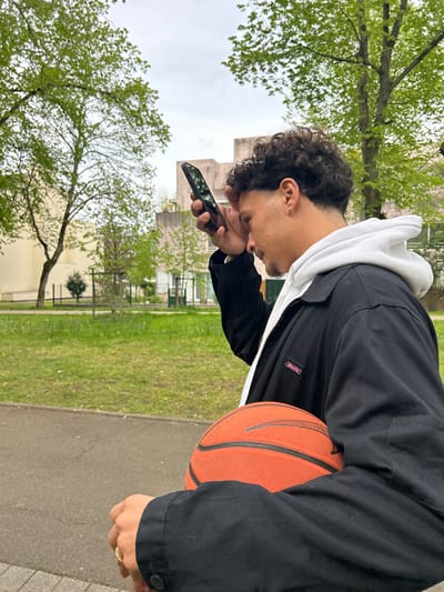 A person stands outdoors on a sidewalk in what appears to be a public park or green space in Noisiel, France. The person is holding a smartphone in their left hand and looking at the screen with their head tilted downwards. They are wearing a black jacket over a white hoodie, and an orange basketball is positioned between their left arm and body. The background includes green grass, trees with budding leaves, and buildings, suggesting an urban environment. The image is oriented in landscape mode. The brand "Dickies" is visible on the person's jacket.