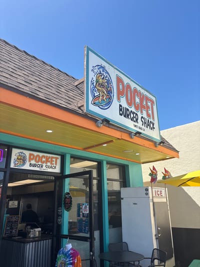 The image shows the "Pocket Burger Shack" restaurant in Sunset Beach, Huntington Beach, California. The restaurant has a large sign with the name on it, plus another sign on the building window. It also has an ice cooler and a table with chairs outside.