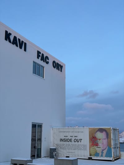 The Kavi Factory building in Henningsvær, Norway, is depicted from an exterior, slightly low-angle perspective under a pale blue sky with faint, scattered clouds. The modern, minimalist structure is predominantly white, featuring the words "KAVI FACTORY" in large, dark, sans-serif lettering affixed to its upper facade. Below the signage, a multi-pane rectangular window is present. At ground level, a glass double door entrance reflects the interior. Snow covers the ground and two low, rectangular concrete blocks in front of the entrance.
Adjacent to the building on the right stands a white shipping container, utilized as a display for a "CONTEMPORARY ART EXHIBITION" titled "INSIDE OUT," with "KAVI FACTORY" branding. The banner includes a list of contributing artists and a prominent graphic of Richard Nixon, whose face is partially obscured by colorful splatters originating from a coffee cup he is holding. A grey cylindrical waste bin with an emblem rests beside the container. In the background, a body of water separates the foreground elements from distant snow-capped mountains under the horizon. No individuals are present.