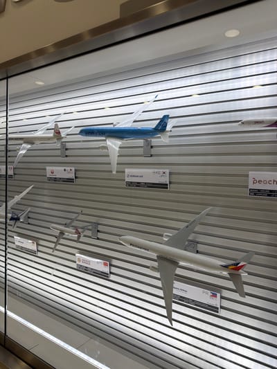 The image depicts a display of airplane models at the ANA Crowne Plaza Hotel Narita in Ota, Japan. The models are mounted on a wall with horizontal silver slats. In the foreground, a model of a Philippine Airlines airplane is prominently displayed. Behind it, a model of a Juneyao Air airplane is visible. Further up, a Korean Air airplane model is shown, along with a Japan Airlines model and a Peach Aviation model. The overall scene is a well-lit display showcasing various airline liveries.