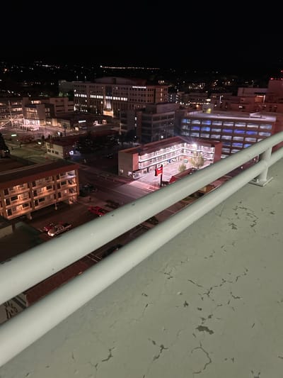 The image displays an elevated nighttime view of downtown Reno, Nevada, United States. In the foreground, a light-colored concrete surface exhibits extensive cracking and wear, partially obscured by a white metal safety railing composed of two horizontal bars. The midground reveals a dense urban environment illuminated by artificial light sources.

To the left, a multi-story motel-style building with a brown tiled roof features visible balconies and a parking area containing three vehicles: a silver/white pickup truck, a dark sedan, and a red pickup truck. A lone individual, appearing as a dark silhouette, is observed walking on a street or sidewalk near this building, heading away from the viewer.

Centrally, a U-shaped motel-style building is prominent, distinguished by a large, red neon sign displaying "INN." Its interior courtyard contains several parked vehicles and two small deciduous trees. Directly behind this structure, a large building with multiple rows of uniformly blue-lit windows is visible, possibly a parking garage.

In the upper-left section of the midground, a prominent multi-story building, clearly identifiable as "Saint Mary's Center for Health," is illuminated. Various streets with multiple vehicles, including a dark pickup truck in motion and a white SUV parked near an intersection, traverse the scene. The background consists of a dark night sky above a sprawling panorama of distant city lights that extend to the horizon, with faint silhouettes of hills or mountains discernible in the furthest distance.
