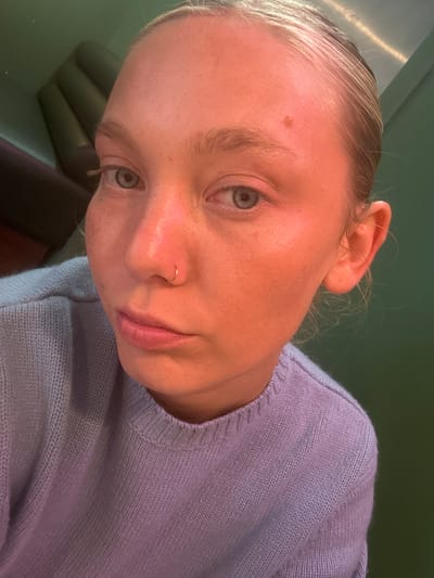 A close-up, angled shot of a fair-skinned person with light blonde hair pulled back from their face, wearing a light purple knitted sweater. The person has light-colored eyes and a small gold nose ring in their left nostril. A small mole is visible on their forehead. The background consists of a light green wall and what appears to be a dark-colored leather or similar material object. The location in Serris, France is not evident from the image.