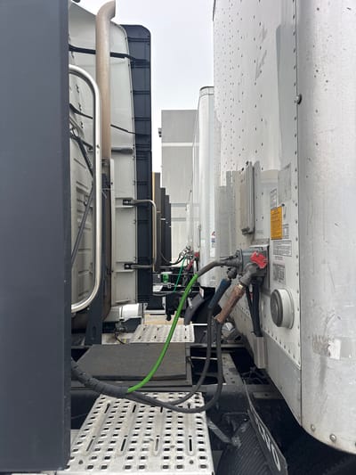 The image captures a close-up perspective of the rear of two semi-trailer trucks parked side-by-side in Santa Clarita, United States of America. The foreground is dominated by the side of a black truck cab on the left, with its ribbed metal platform and railing visible. To the right, the silver, riveted side of a trailer is prominent, featuring various connectors and cables, including a green air hose and red and blue electrical connectors, attached to a coupling assembly. Several warning labels are affixed to the trailer's surface. In the background, other semi-trailer trucks are visible, lined up and facing the same direction, suggesting a truck stop or depot. The sky is overcast, indicating a potentially cloudy or hazy day. The overall scene is industrial, focusing on the mechanical details of commercial vehicles.