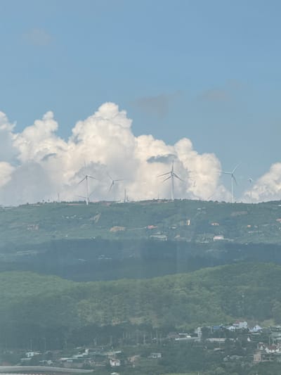 The image depicts a landscape with a clear blue sky and scattered white clouds, above rolling green hills. Several wind turbines are visible on the crest of the hills, with their blades spread. In the foreground and further down the hills, buildings and houses are scattered amongst the greenery, suggesting a settlement. The overall scene appears to be a rural or semi-rural area with evidence of wind energy infrastructure. The location is Đa Lộc, Vietnam.