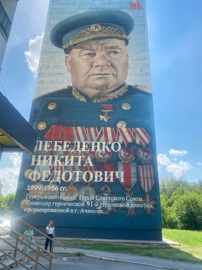 The image shows a large mural painted on the side of a building, depicting a portrait of a man in a military uniform. The man is wearing a dark blue cap with a red star and gold trim, and a dark blue uniform with gold epaulets and numerous medals. The mural is set against a backdrop of blue sky with white clouds and green trees.
Below the portrait, text in white letters identifies the subject as "ЛЕБЕДЕНКО НИКИТА ФЕДОТОВИЧ". Further down, the text reads "1899-1956 гг." and "Генерал-лейтенант, Герой Советского Союза, Командир героической 91-й стрелковой дивизии, сформированной в г. Ачинске." This translates to "Lieutenant General, Hero of the Soviet Union, Commander of the heroic 91st Rifle Division, formed in the city of Achinsk." This indicates the mural is located in Achinsk, Russia.
Two women are visible in the foreground, walking up a set of outdoor stairs leading to the building. One woman is wearing a light green dress, and the other is wearing a white t-shirt and dark pants. They appear to be interacting with each other. The building itself has a white facade and a dark awning extending from its side. The environment appears to be an urban setting with a street visible in the lower left corner. The time of day appears to be daytime, with bright sunlight casting shadows.