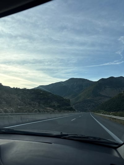 The image depicts a view from inside a vehicle traveling on a highway that cuts through a mountainous, possibly rural, area. The highway has two lanes in each direction, marked with solid white lines. A concrete barrier separates the lanes, and a guardrail is visible on the right side of the road. The surrounding landscape is characterized by rolling hills and mountains covered in vegetation, with a clear blue sky filled with wispy white clouds above. The lighting suggests it is daytime, possibly late afternoon, with the sun casting shadows on the mountains. There are no visible people or other vehicles in the immediate vicinity. The interior of the vehicle, including the dashboard and windshield wipers, is visible in the foreground, indicating the photo was taken from the driver's perspective. The location is specified as Γυμνότοπος, Greece.