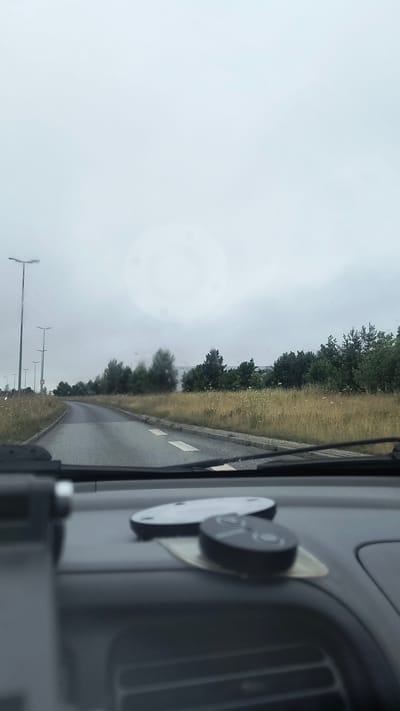 This is a first-person perspective photograph taken from inside a vehicle, likely a bus or truck, looking out the side window. The scene is oriented vertically. The dominant elements are the road, vegetation, and sky. A paved road curves gently to the left, bordered by a dry, grassy verge with scattered trees. The sky is overcast, suggesting a cloudy or possibly rainy day. In the foreground, within the vehicle's interior, a dark grey dashboard is visible on the right side of the frame. Attached to the dashboard are two circular objects, one black and one white, possibly a GPS device or phone mount. The overall mood is subdued, consistent with the overcast weather. The location is identified as Ølby, Denmark. There is no visible text in the image. The image captures a moment of travel, likely during daylight hours, given the brightness of the sky, though the cloud cover obscures direct sunlight.