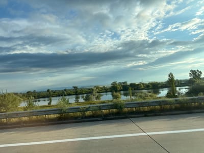 The image captures a scenic view of a riverbank lined with trees, seen from the perspective of a moving vehicle on a highway. The foreground shows a concrete road with a white line marking the lane. A metal guardrail separates the road from the natural landscape. Beyond the guardrail, a calm river reflects the sky, which is filled with dramatic clouds. The river is bordered by lush green vegetation and scattered trees. In the distance, rolling hills are visible under the expansive sky. The overall atmosphere is tranquil and serene, suggesting a rural or semi-rural setting. The location is identified as Samtsevrisi, Georgia. There are no people or explicit actions depicted in the image.