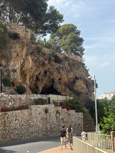 The image depicts a rocky cliff face with a cave opening, nestled amidst lush greenery and terraced stone walls. Several individuals are visible on a paved walkway adjacent to the cliff, suggesting a public area or tourist attraction. One person, wearing a white shirt and patterned shorts, leans on a light-colored railing. Further down, two more people are walking, one with a backpack. The setting appears to be outdoors during the daytime, with bright sunlight casting shadows and illuminating the scene. The sky is partly cloudy, indicating fair weather.

The stone walls are constructed with rough, irregular stones, creating a textured and rustic appearance. Red flowers are planted along one of the terraces, adding a splash of color to the predominantly earthy tones of the rock and stone. The vegetation includes trees with dense foliage, some of which appear to be pine trees, and palm trees. A white street lamp is mounted on a pole extending over the cliff, and a modern-looking white building with multiple windows is visible in the upper right background, hinting at an urban or resort environment. The road is visible on the right side of the image, with a red and white traffic cone on it.

Given the location context of Benalmádena, Spain, this scene likely represents a natural feature integrated with man-made structures in a tourist-oriented area. The overall impression is one of a scenic and accessible natural wonder, possibly a tourist spot with walking paths and viewpoints. No discernible text is visible in the image.