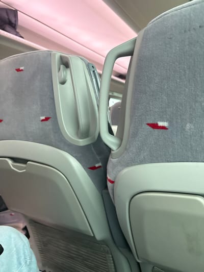 The image shows the interior of a bus. The back of a seat is visible, with gray fabric and a red and white design. The seat is reclined and the back of the seat in front is visible. There is a handle on the back of the seat. The seats appear to be in good condition. The bus interior is clean and well-maintained. There is a small white storage compartment behind the seat. The image is taken from a low angle, looking up towards the seat. The coordinates provided place the image in Otero de Herreros, Spain.  However, the image only shows the interior of a bus, and there is no way to determine if this bus is in Otero de Herreros.