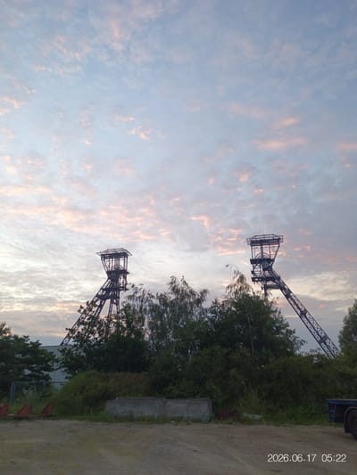 The image depicts two industrial headframes, likely remnants of a former coal mine, silhouetted against a sky filled with scattered clouds tinged with pink and orange from what appears to be either sunrise or sunset. The headframes are tall, lattice-like metal structures. In the foreground, there is overgrown vegetation, including trees and bushes. The ground in front of the vegetation is a rough, unpaved surface. In the lower right corner, a date and time stamp reads "2026.06.17 05:22". Based on the provided metadata, the scene is located in Houthalen, Belgium.