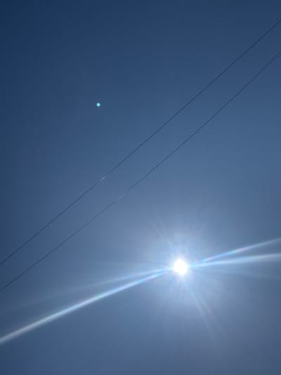 The image shows a clear blue sky with two power lines running diagonally from the top left to the bottom right. The sun is a bright, star-like object in the bottom right quadrant of the image, casting a lens flare that creates a rainbow effect. There is a small, circular, bright blue object in the upper left quadrant of the sky, possibly a reflection or another celestial body. The scene is devoid of any people or specific landmarks that would indicate a location in Muravlenko, Russia.
