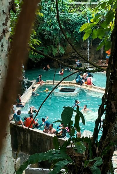 The image depicts a natural swimming pool in a lush, verdant environment, likely in Penungkiren, Indonesia. The water is a vibrant turquoise, suggesting it is clean and possibly fed by a waterfall or spring, as suggested by a small cascade visible on the rocky cliff face in the background.
A diverse group of people, appearing to be locals and tourists, are enjoying the water and the surrounding area. Several individuals are swimming or wading in the pool. Some are seated on the edge of the pool, which is constructed with concrete walls, while others are floating or interacting in the water. Children are visible among the swimmers, some wearing life vests.
The environment is characterized by dense foliage, with large green leaves and tree trunks framing the scene, suggesting a tropical or subtropical setting. The rocky, textured cliff face in the background is also covered in vegetation, contributing to the natural, almost secluded atmosphere. The presence of a central, square structure within the pool, possibly a submerged platform or fountain, adds an architectural element to the natural landscape.
The overall impression is one of a recreational area where people are relaxing and engaging in water activities within a beautiful, natural setting.