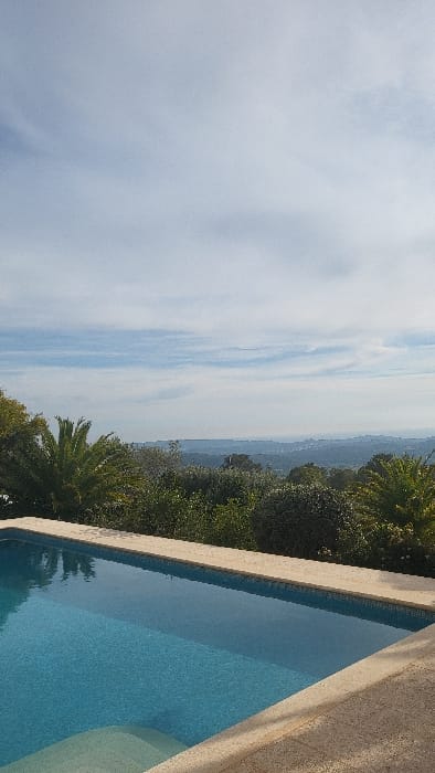 The scene is an exterior view, likely from a residential or leisure property, within or overlooking the Polígon Industrial De Calvià, Spain. A rectangular swimming pool, filled with clear blue water and featuring visible blue mosaic tiling at its bottom, occupies the lower portion of the frame. Light-colored, textured coping stones surround the visible edges of the pool, with integral steps evident at the lower-left corner. Beyond the pool, a band of dense green Mediterranean vegetation, including prominent palm trees, forms a natural barrier across the midground. In the distance, an expansive landscape of rolling hills with scattered urban development is visible, extending towards a hazy horizon line. The upper half of the image is dominated by a light blue sky with scattered, thin white clouds. No human figures, animals, or active subjects are depicted. The environment is illuminated by natural daylight, indicating clear weather conditions.
