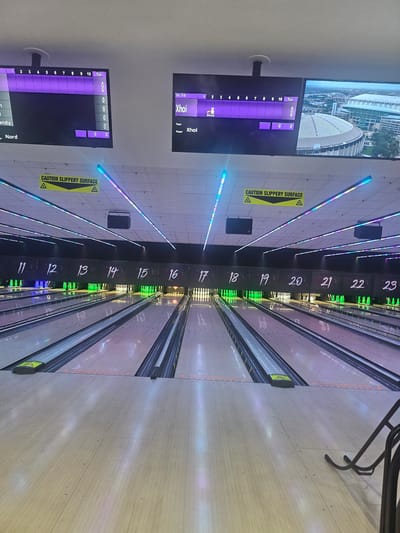 An indoor bowling alley is depicted, viewed from the perspective of a bowler facing the lanes. The lanes are numbered sequentially from 11 to 23, with bowling pins set up on several of them. Overhead, digital scoreboards display game information. One scoreboard shows "Fr.70 Xhoi" under "Team" and "Player" respectively, with a score of "0" in the total column. Another scoreboard to the left, partially obscured, shows a score of "0" for a player named "Nord". Yellow caution signs indicating a "SLIPPERY SURFACE" are visible on the ceiling. LED strip lighting in various colors illuminates the bowling alley. The overall environment is dark with brightly lit lanes and scoreboards. There is no indication of the city of Gloucester, United Kingdom within the image.