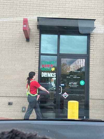 Here is a description of the image:
The image shows the exterior of a building, likely a restaurant in Mountain Park, United States of America. The building has a brick facade with a dark-framed glass door. On the door are several signs, including one advertising "Strawberry Hibiscus Drinks," another displaying the restaurant's operating hours (6:00 AM to 9:00 PM for dining room and 6:00 AM to 10:00 PM for drive-thru, Monday to Saturday) and indicating it's closed on Sundays. Other signs state "Smoke Free Property" and "No Soliciting."
A person wearing a red shirt and a safety vest is visible in front of the building. The person is facing away from the camera. A red box is fixed on the wall of the building to the left of the door. An electrical outlet or similar fixture can be seen on the wall to the right of the door, above a possible security camera.
The overall impression is that it's a daytime scene at a fast-food or casual dining establishment.
