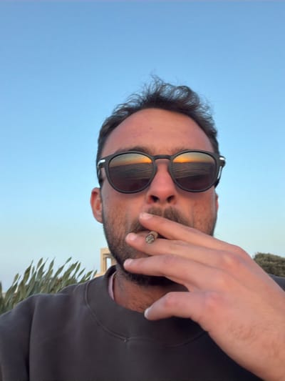 An adult male is depicted in a close-up, slightly low-angle frame, presenting a self-portrait. He possesses dark, short, curly hair, a dark beard, and a mustache. He wears black-framed sunglasses with reflective lenses, which distinctly show a sunset over a body of water, displaying an orange sun low on the horizon and visible waves. The male is dressed in a dark grey crewneck sweatshirt over a light pink collared shirt. His right hand is raised, holding a lit, partially consumed cigarette or cigarillo between his index and middle fingers, positioned near his mouth. The cigarette/cigarillo tip exhibits ash and a glowing ember. The background consists of a clear, light blue sky. To the subject's left, out-of-focus green, spiky foliage is visible. To the right, a light-colored, out-of-focus structure or building is present. The scene occurs at sunset in a coastal environment in the city of Beit Yanai, Israel.