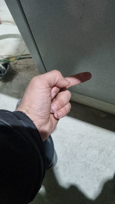 The image depicts a close-up, angled view of a human left hand extending its index finger, with the remaining fingers and thumb curled into the palm. The fingernails show visible dirt or grime, and the hand appears to be that of an adult. The subject is wearing a dark-colored sleeve, likely part of a jacket or sweater, and a portion of blue legwear is visible below. The extended index finger casts a distinct shadow onto a large, grey, textured vertical surface that occupies the right side and upper portion of the frame. This surface exhibits a subtle vertical seam on its left edge. The floor, visible in the lower-left, appears to be light-colored concrete or a similar material. In the far background on the left, an indistinct area shows what appears to be wires (green and yellow visible) and a dark, possibly wheeled, object, suggesting an indoor or industrial-type setting. Strong directional lighting creates clear shadows. This scene is located in Chapyk, Uzbekistan.