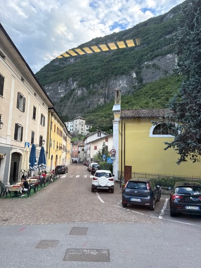 The image depicts a street scene in Mezzolombardo, Italy, likely during daylight hours. The street is paved with cobblestones and lined with buildings on both sides. The architecture suggests a European town, with multi-story buildings featuring traditional windows and shutters. On the left, a building with a light-colored facade has tables and chairs set up outside, indicating a cafe or restaurant, with several patrons seated. Blue and white umbrellas provide shade over the outdoor seating.

The street slopes gently upwards into the distance. Several cars are parked along the right side of the street and some are in motion. A white SUV is parked on the right, followed by a dark gray hatchback and a black sedan. Further up the street, more parked cars are visible.

In the background, a large, tree-covered mountain rises steeply. The sunlight creates a pattern of rectangular shapes, possibly due to reflections or sunlight filtering through something, on the upper part of the mountain. A yellow building with a tiled roof and a tall, slender bell tower is prominent on the right side of the street. A few traffic signs are visible, including a pedestrian crossing sign and a no-trucks sign. The sky is partly cloudy, with patches of blue visible.