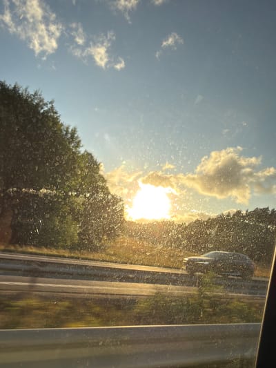 The image captures a sunset view from a vehicle traveling on a highway in Ödsmål, Sweden. The sun is low on the horizon, casting a golden glow through scattered clouds. Trees line the sides of the highway, their foliage silhouetted against the bright sky. A car is visible driving on the adjacent lane, its form blurred by motion. The foreground shows a portion of the vehicle's interior, including the window, which is speckled with what appears to be water droplets or dirt. The overall scene conveys a sense of movement and the serene beauty of a twilight drive.