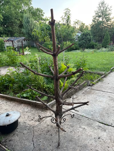The image depicts a backyard scene in Yenakiieve, Ukraine, dominated by a rustic, bare tree-like structure with branches extending outwards, likely intended as a decorative element or for holding items. The structure stands on a metal base with decorative scrollwork. It is positioned on a cracked concrete patio. To the left of the structure, a large, lidded metal pot is visible. The background reveals a garden with lush green grass, various plants, including lilies and ferns, and a small wooden gazebo. In the distance, trees and a residential building are discernible, suggesting a suburban or rural setting. The overall impression is one of a lived-in, somewhat overgrown garden space.