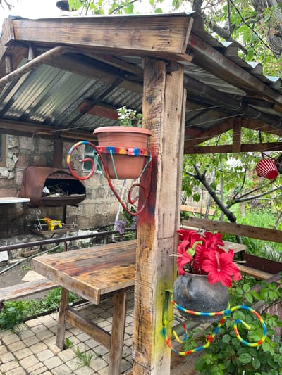 In Lusagyugh, Armenia, a rustic outdoor structure, likely a gazebo or covered seating area, is depicted. The structure is primarily constructed from weathered wood and corrugated metal roofing. A wooden table and benches are set on a paved patio area. Various decorative elements are present, including potted plants, a metal grill, a toy wheelbarrow, and brightly colored accents spray-painted onto the wooden supports. In the foreground, a pot of red flowers is held by a decorative metal stand. The surrounding environment is lush with green foliage and trees, suggesting a garden or yard setting. The overall impression is one of a handmade, perhaps somewhat cluttered, but functional outdoor space.