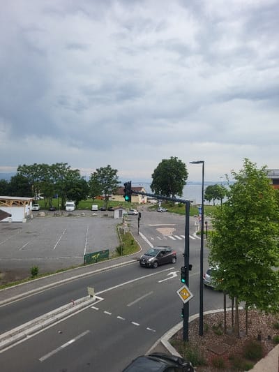 The image is taken from a high vantage point looking down at a road intersection in the town of Publier, France, located at latitude 46.394 and longitude 6.54. The intersection has a traffic light at the center of the image, with a green light for the cars traveling towards the right side of the image. A grey sedan is traveling towards the right, having just navigated the intersection. To the left is a parking lot with trees and some cars. There is a small building on the left side of the parking lot. There is another car driving on the right side of the intersection, and there is a car parked on the right side of the intersection just beyond the traffic lights. The scene is captured under a cloudy sky. The scene is shot from a high vantage point, likely from a building overlooking the intersection. The weather is overcast, with a gray sky.