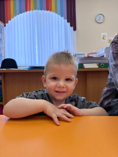 A young child with short, light-colored hair leans against an orange table. The child wears a gray shirt with a darker pattern. The background includes a window with colorful slats, a wall clock, and shelves with papers and files. The location in the city of Yaroslavl', Russia, cannot be verified based solely on this image.