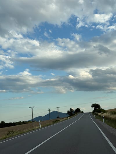The image shows a view of a road in Luhyňa, Slovakia, leading into a gently sloping hill. The road is paved with asphalt and has white lines marking the lanes. The sky is partly cloudy with a mix of blue sky and grey and white clouds. On either side of the road, there are fields of dry grass and some scattered trees. In the distance, there are mountains and power lines. There is a single car visible on the road in the far distance, its headlights on. There are no people visible in the image. The scene appears to be a rural landscape.